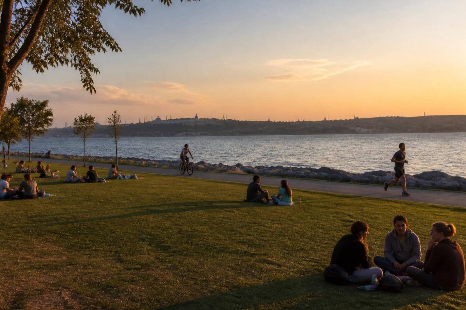 People sitting on lawns and rocks beside a calm sea at sunset, with a faint city skyline across the water