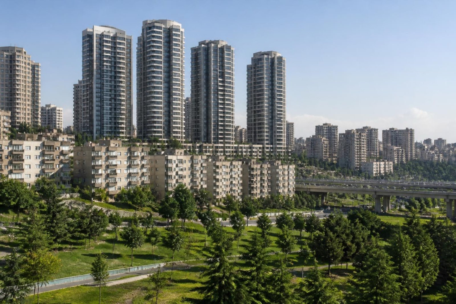 Bir yamaçta eski orta katlı apartman bloklarının yanında yükselen yeni konut kuleleri; eteklerinde yeşil bir park